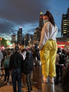 Student standing above a crowd of protesters at dusk wearing mask, looking back at the camera